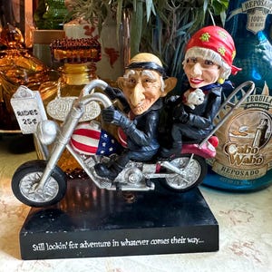 May include: A figurine of two elderly characters riding a motorcycle with a red, white, and blue fuel tank. The motorcycle has a sign reading RIDES 25¢. The black base features the text: Still lookin' for adventure in whatever comes their way...