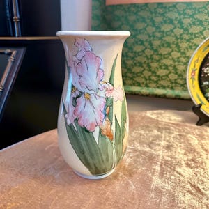 May include: A beige ceramic vase with a hand-painted design of pink and white iris flowers and green leaves. The vase has a flared top and a rounded body. The floral design is detailed and artistic.