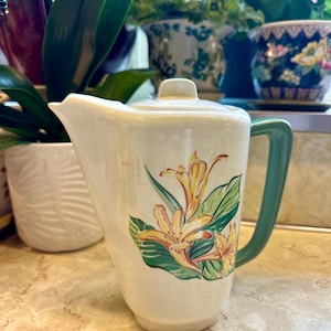 May include: Cream-colored ceramic pitcher with a green handle and lid. The pitcher features a floral design with yellow flowers and green leaves. The pitcher is a vintage style and is likely used for serving beverages.