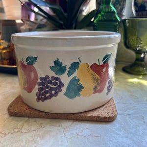 May include: A cream-colored ceramic crock with a fruit design featuring apples, pears, and grapes. The crock sits on a square cork coaster. The background includes other decorative glass items.