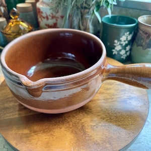 May include: A round, brown ceramic saucepan with a single handle and a small pouring spout. The exterior features a glossy, two-toned glaze with a darker brown upper section and a lighter, earthy brown base, showcasing a smooth, reflective finish.