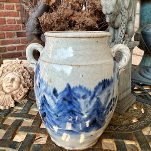May include: A ceramic vessel featuring a cream-colored base with a prominent crackle glaze finish. The exterior is decorated with a blue painted landscape depicting a forest of pine trees. It includes two side handles and a wide circular opening.