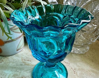 May include: A vibrant turquoise blue glass compote dish featuring a ruffled, scalloped rim and a pressed pattern of circular motifs and starbursts on the bowl and circular base. The glass has a translucent quality with a polished finish.