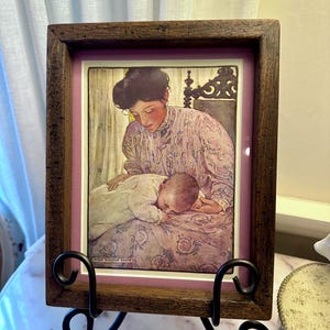 May include: A framed print of a mother and child in a vintage style. The print is set in a dark brown wooden frame with a pink mat. The print depicts a woman in a floral dress and a baby. The frame is displayed on a black metal stand.