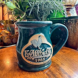 May include: A dark teal ceramic mug featuring a raised beige emblem with a rooster illustration. The text reads Egg Harbor Cafe, Breakfast & Lunch, 1985, Lake Forest, Illinois. The mug has a wide handle and a smooth, glossy finish throughout.