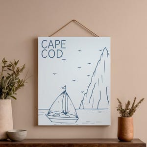 Può includere: Stampa su tela incorniciata con un'illustrazione minimalista di una barca a vela, una montagna e uccelli, con la scritta "CAPE COD" in blu navy. L'opera è sospesa da un sottile cordino marrone.