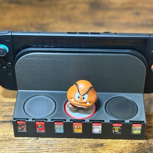 May include: Black Nintendo Switch console resting on a black stand with game cartridge slots. A small orange Goomba figure is centered on the stand. Several game cartridges are visible in the slots.