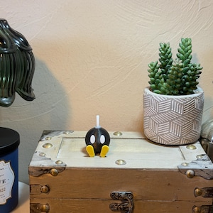 May include: A black and white cartoon bomb figurine with yellow feet and a white fuse sits on a wooden chest. A small succulent plant in a patterned pot and a decorative glass bottle are also visible.