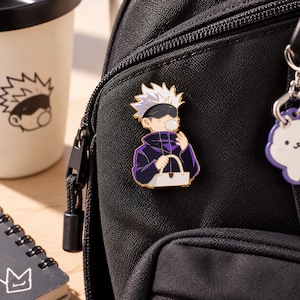 Peut inclure: Une broche en émail métallique représentant un personnage de dessin animé aux cheveux blancs en pointe, portant un bandeau noir et une veste violette à col haut. Le personnage fait une bulle de chewing-gum et tient un sac blanc sur un sac à dos.