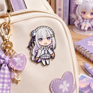 May include: A metal enamel pin featuring a chibi-style anime character with long silver hair and purple eyes, wearing a white and purple outfit. The pin is attached to a cream-coloured bag adorned with a purple heart patch and a dangling heart charm.