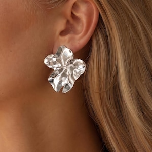 May include: A silver earring with an organic, irregular, and wavy petal-like shape. The surface of the earring is polished and reflective, catching light with its undulating, fluid contours that resemble a blooming flower or molten metal form.