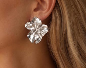 May include: A silver earring with an organic, irregular, and wavy petal-like shape. The surface of the earring is polished and reflective, catching light with its undulating, fluid contours that resemble a blooming flower or molten metal form.
