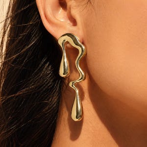 May include: Gold-coloured, abstract earrings with a flowing, organic design. The earrings have a polished finish and a dangling teardrop shape at the bottom. The jewellery is worn on the ear.