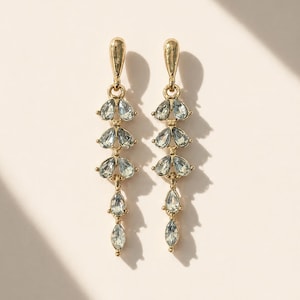 Peut inclure: Une paire de boucles d'oreilles pendantes dorées. Chaque boucle d'oreille présente une série de cristaux bleus clairs en forme de goutte d'eau, tombant d'un dessus décoratif. Les cristaux sont disposés en motif floral superposé.