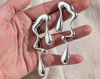 May include: A pair of silver-toned earrings featuring an elongated, fluid, and abstract shape that resembles dripping liquid. The earrings have a smooth, reflective, and polished metallic surface with curved, organic lines and a teardrop-like base.