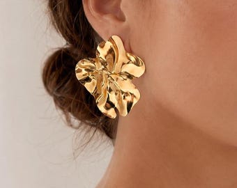 May include: Gold-coloured, flower-shaped earrings with a textured, hammered finish. The earrings have a large, statement design, with multiple petals and a shiny appearance. The jewellery is worn on the ear, and the image is a close-up of the earrings.