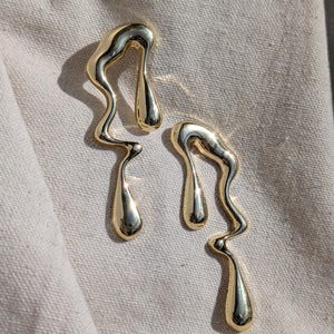 May include: A pair of gold-toned earrings featuring an abstract, fluid, and organic shape that resembles melting liquid. Each earring has a smooth, polished, and reflective surface with elongated, curved lines and a teardrop-shaped end.