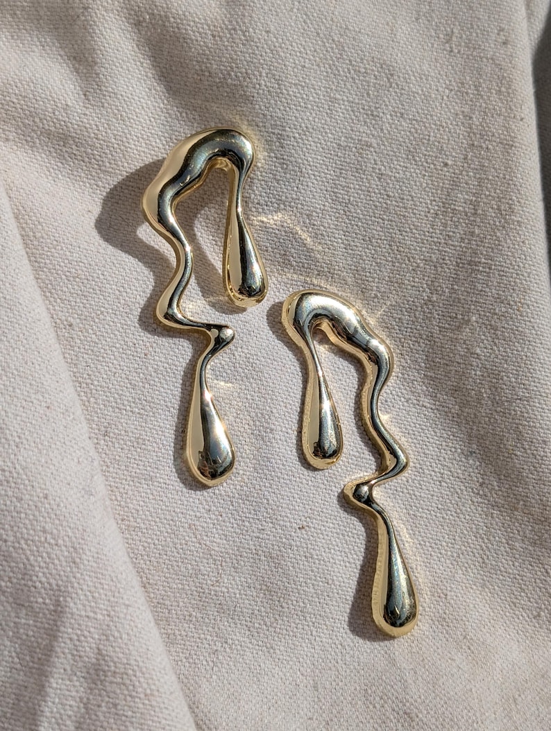 May include: A pair of gold-toned earrings featuring an abstract, fluid, and organic shape that resembles melting liquid. Each earring has a smooth, polished, and reflective surface with elongated, curved lines and a teardrop-shaped end.