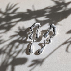 May include: A pair of silver-toned earrings featuring an irregular, fluid, and melted metal design. Each earring has a long, wavy, and organic shape with smooth, reflective surfaces that catch the light, creating a liquid-like appearance.
