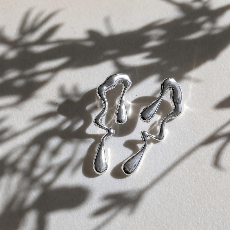 May include: A pair of silver-toned earrings featuring an irregular, fluid, and melted metal design. Each earring has a long, wavy, and organic shape with smooth, reflective surfaces that catch the light, creating a liquid-like appearance.