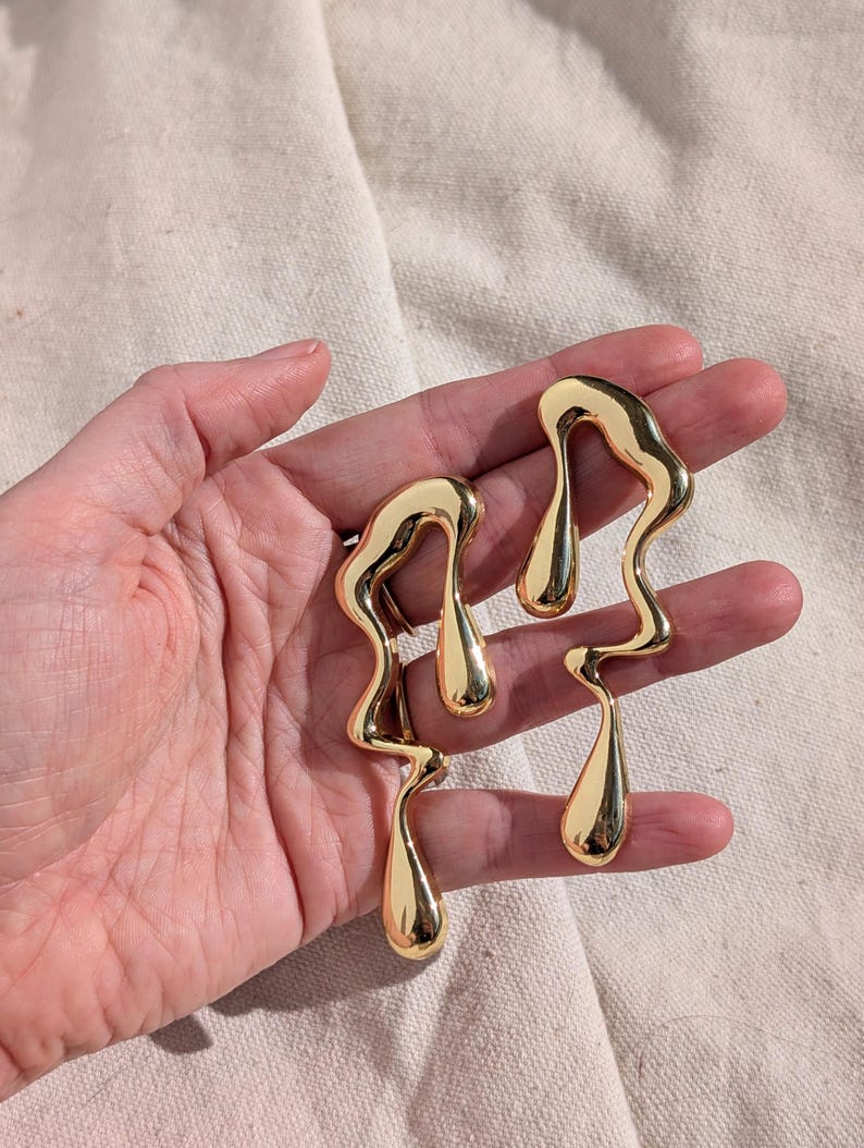 May include: A pair of gold-toned earrings featuring an elongated, fluid, and abstract molten metal shape. The design consists of smooth, reflective, and irregular curves that taper into teardrop-like ends, creating a continuous, organic silhouette.