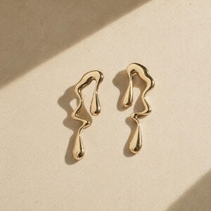 May include: A pair of gold-toned earrings featuring an irregular, fluid, molten metal design. Each earring has a wavy, elongated silhouette with a smooth, reflective surface and a teardrop-shaped element at the bottom, creating a liquid-like appearance.