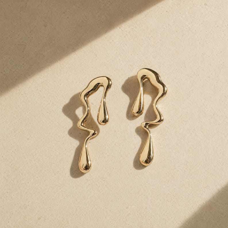 May include: A pair of gold-toned earrings featuring an irregular, fluid, molten metal design. Each earring has a wavy, elongated silhouette with a smooth, reflective surface and a teardrop-shaped element at the bottom, creating a liquid-like appearance.