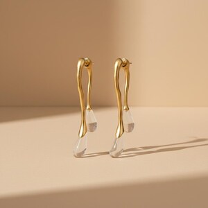 May include: A pair of gold-toned drop earrings featuring a fluid, elongated metal frame with a matte finish. Each earring suspends two clear, teardrop-shaped acrylic or glass pendants that hang at varying lengths from the curved metal structure.