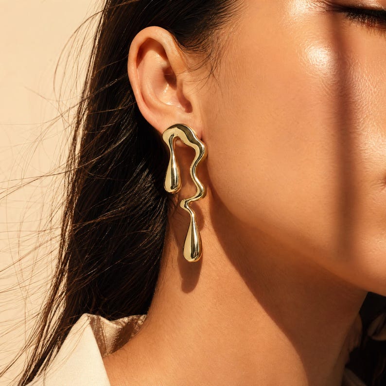 May include: A gold-toned earring featuring an elongated, fluid, and organic shape that resembles a melting or dripping form. The metal surface is smooth and highly reflective, catching the light with a polished finish that hangs from the earlobe.