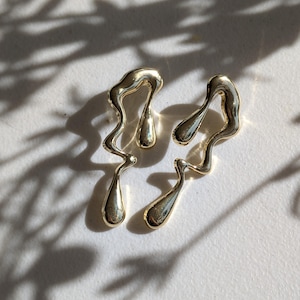 May include: A pair of gold-toned earrings featuring an irregular, fluid, and melted metal design. Each earring consists of a wavy, elongated frame with two smooth, teardrop-shaped accents integrated into the structure, creating a liquid appearance.