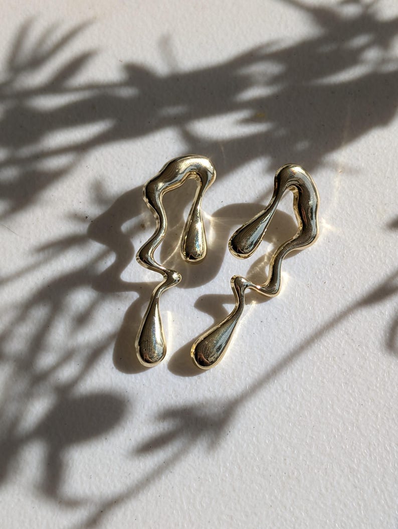 May include: A pair of gold-toned earrings featuring an irregular, fluid, and melted metal design. Each earring consists of a wavy, elongated frame with two smooth, teardrop-shaped accents integrated into the structure, creating a liquid appearance.