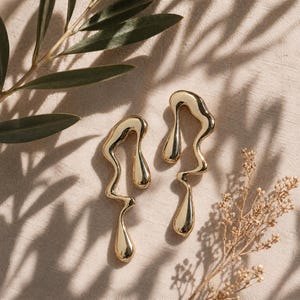 May include: A pair of gold-toned earrings featuring an elongated, fluid, and irregular molten metal shape. Each earring has a smooth, reflective surface with a continuous, winding design that terminates in a teardrop-like form at the bottom.