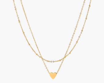 May include: A gold-colored, layered necklace with a heart pendant. The necklace features two delicate chains, one with small bead accents. The heart pendant is smooth and centered, creating a simple yet elegant design.