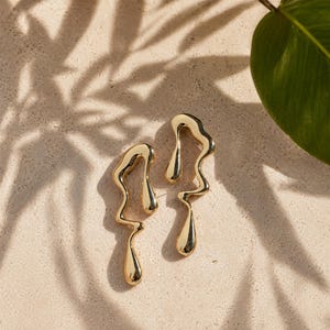 May include: A pair of gold-toned earrings featuring an irregular, fluid, and molten-like shape. Each earring has a smooth, reflective surface with a curved, elongated design that tapers into a teardrop form at the bottom, creating a distinct metallic aesthetic.