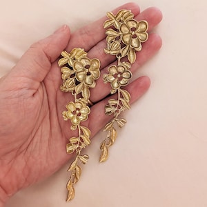 May include: Gold-toned floral earrings with a cascading design. Each earring features multiple flower and leaf clusters, creating a delicate, botanical aesthetic. The earrings are held in a hand, showcasing their intricate details and elegant style.