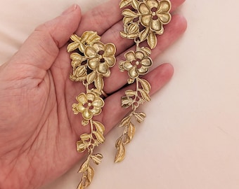 May include: Gold-toned floral earrings with a cascading design. Each earring features multiple flower and leaf clusters, creating a delicate, botanical aesthetic. The earrings are held in a hand, showcasing their intricate details and elegant style.