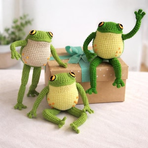 Puede incluir: Tres juguetes de rana hechos a mano con ganchillo en varias poses. Las ranas tienen cabezas y extremidades verdes, vientres amarillos y ojos grandes y redondos. Están sentadas sobre y alrededor de una caja de regalo marrón con una cinta turquesa.