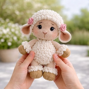 Puede incluir: Una muñeca de oveja de ganchillo hecha a mano con un cuerpo blanco esponjoso, cara beige y pezuñas marrones. La oveja tiene grandes ojos negros, mejillas rosadas y pequeños acentos de flores rosas. La muñeca está en las manos de una persona.