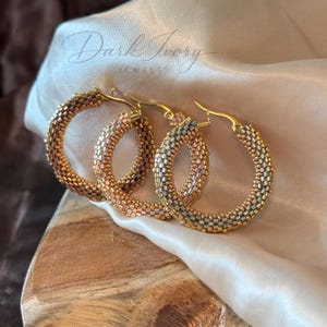 Puede incluir: Un par de pendientes de aro con un patrón de cuentas tejidas en tonos metálicos de oro, bronce y plata. Los aros circulares están construidos con pequeñas cuentas uniformes e incluyen un mecanismo de cierre de metal dorado en la parte superior.