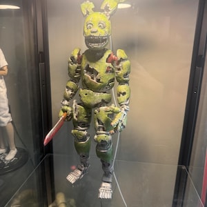 May include: A weathered, olive-green animatronic rabbit figure with a distressed, damaged exterior. The figure features a wide, toothy grin, large eyes, and articulated limbs. It holds a small, silver-colored knife with a brown handle in its right hand.
