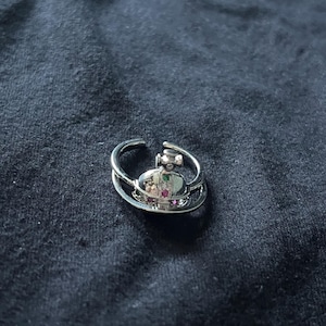 Könnte beinhalten: Ein silberfarbener Ring mit einem zentralen Kugel-Emblem, das von einem dünnen kreisförmigen Band umgeben ist. Die Kugel ist mit kleinen, mehrfarbigen Steinen besetzt, darunter rote und grüne Akzente, die in einen polierten Metallrahmen gefasst sind.