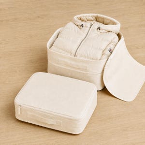 May include: A cream-colored quilted hooded jacket with a front zipper and a small circular logo on the sleeve, stored inside an open cream-colored fabric storage box with a matching rectangular lid placed separately in the foreground.