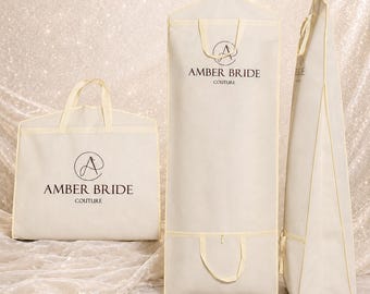 May include: Three off-white garment bags with cream trim and handles. One square bag and two tall rectangular bags are displayed. Each bag features the black printed logo and text AMBER BRIDE COUTURE. The bags are made of a non-woven fabric material.