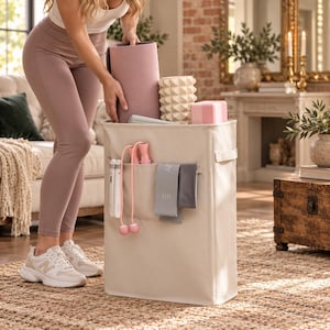 May include: A tall, rectangular, beige fabric storage bin featuring exterior pockets. The pockets contain a white jump rope with pink handles, a pink jump rope, and two gray resistance bands. The bin is filled with a rolled mauve yoga mat.