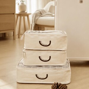 May include: A set of three rectangular cream-colored fabric packing cubes stacked vertically. Each cube features a dark brown curved line graphic with the text "Nice to meet you" printed below it and a zipper closure along the top edge.