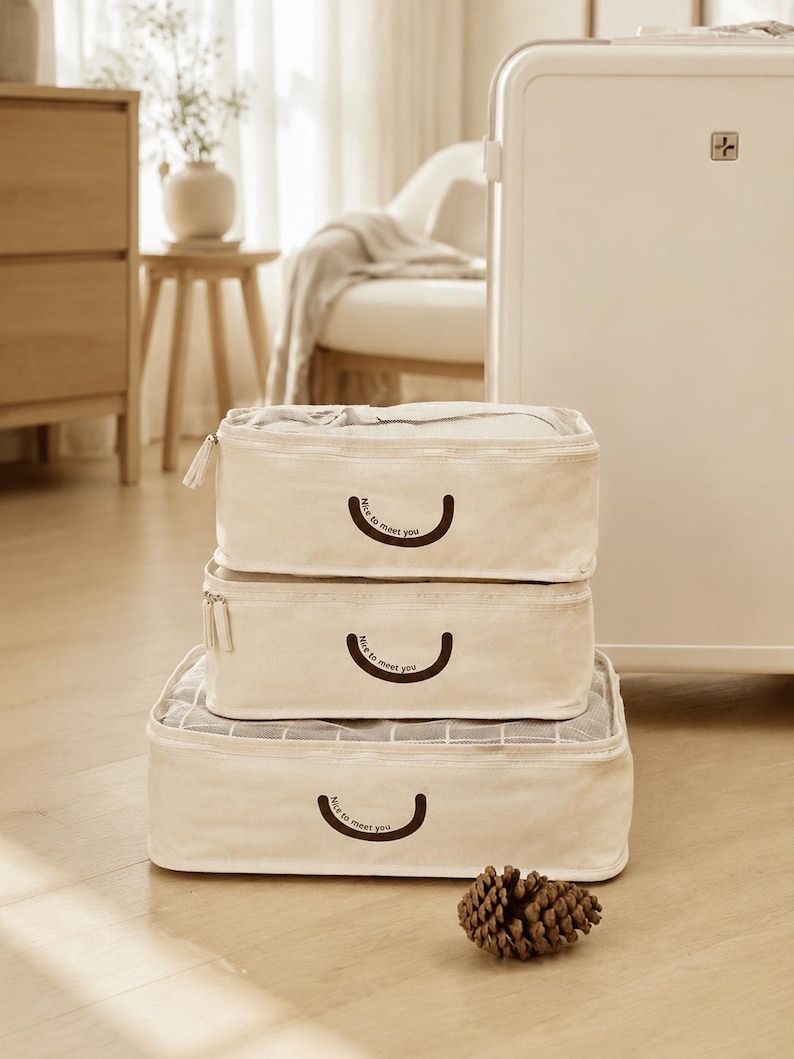 May include: A set of three rectangular cream-colored fabric packing cubes stacked vertically. Each cube features a dark brown curved line graphic with the text "Nice to meet you" printed below it and a zipper closure along the top edge.
