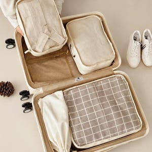 May include: An open beige suitcase containing four beige packing organizers. One organizer holds a folded light-colored shirt, another is plain, one features a white grid pattern on a gray background, and one is a drawstring pouch. The suitcase has wheels.