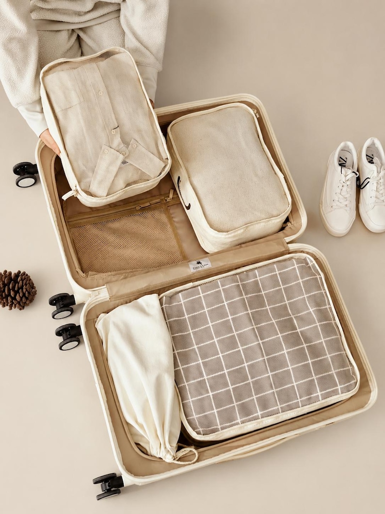 May include: An open beige suitcase containing four beige packing organizers. One organizer holds a folded light-colored shirt, another is plain, one features a white grid pattern on a gray background, and one is a drawstring pouch. The suitcase has wheels.