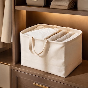 Op de afbeelding: Een rechthoekige off-white canvas opbergmand met twee stoffen handvatten aan de voorzijde. De mand is gevuld met netjes opgevouwen lichtgekleurde kledingstukken, waaronder witte en beige kleding, georganiseerd in het open bovenste compartiment.