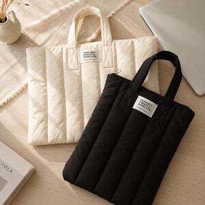 May include: A cream-coloured and a black quilted laptop sleeve, each featuring two top handles, a zipper closure, and a rectangular white fabric label with the text YUAN BEN CREATE A GOOD LIFE. Going more simply printed in black ink.
